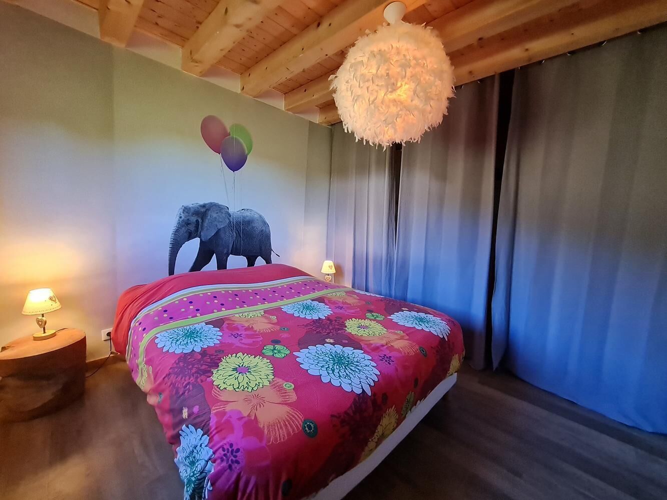 Une chambre avec un plafond en bois, une lampe blanche et moelleuse, un lit recouvert d’une courtepointe à motifs floraux colorés et une décoration murale représentant un éléphant.