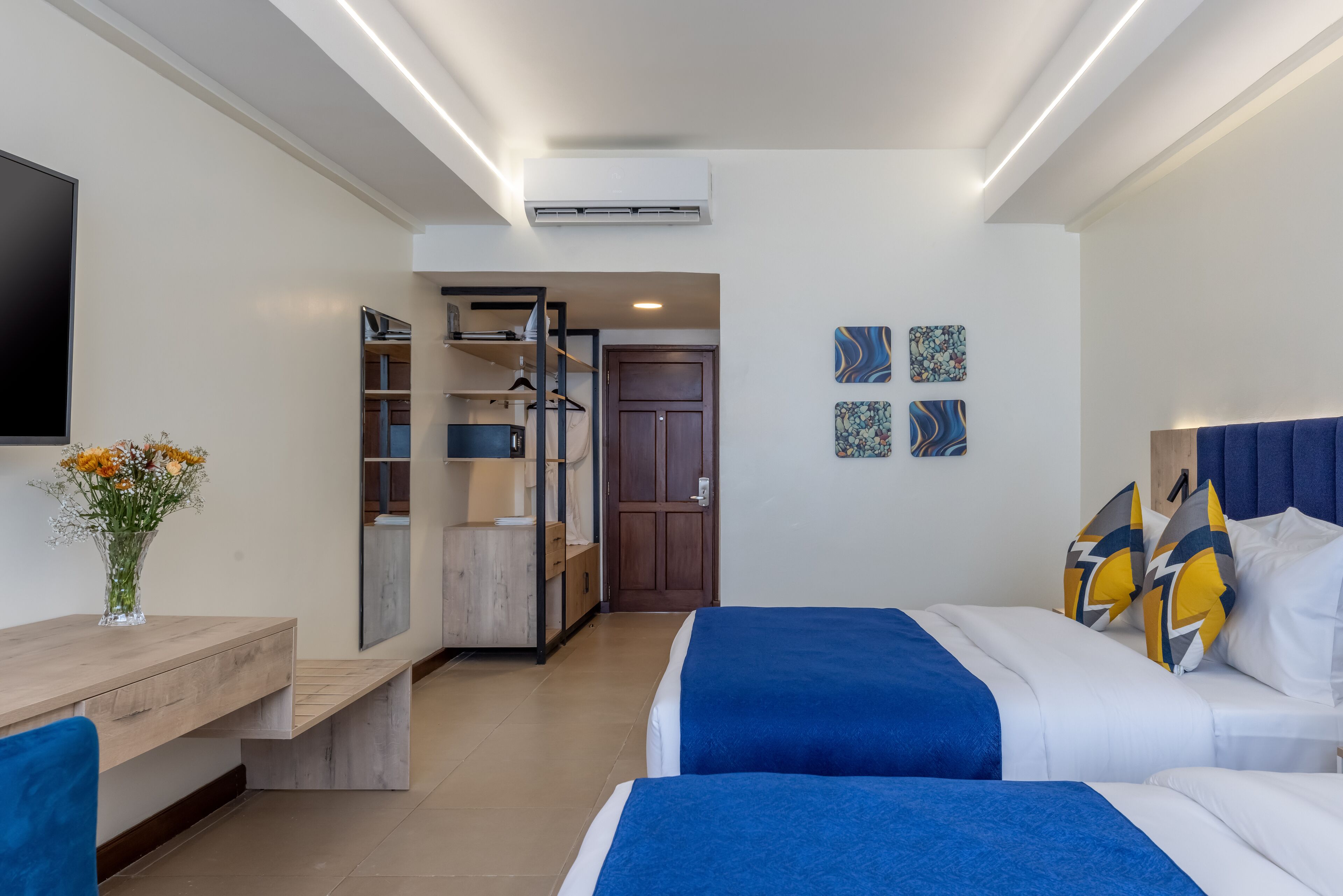 Una habitación de hotel con cama, mesita de noche y escritorio con silla. Se observa un armario, un espejo y una puerta que da a otra habitación.