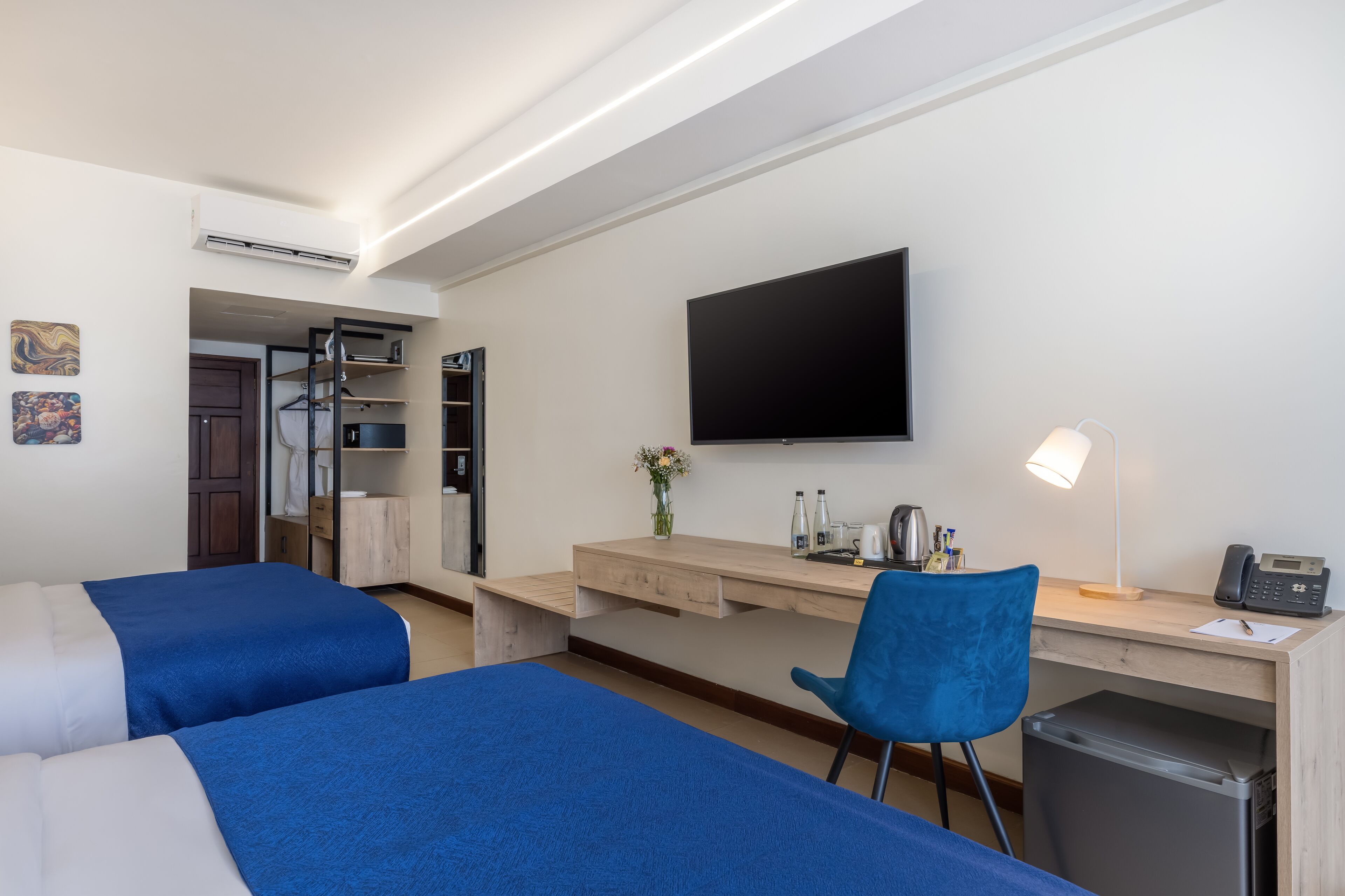 A hotel room with two beds, a desk, a chair, a TV, and a small kitchenette.