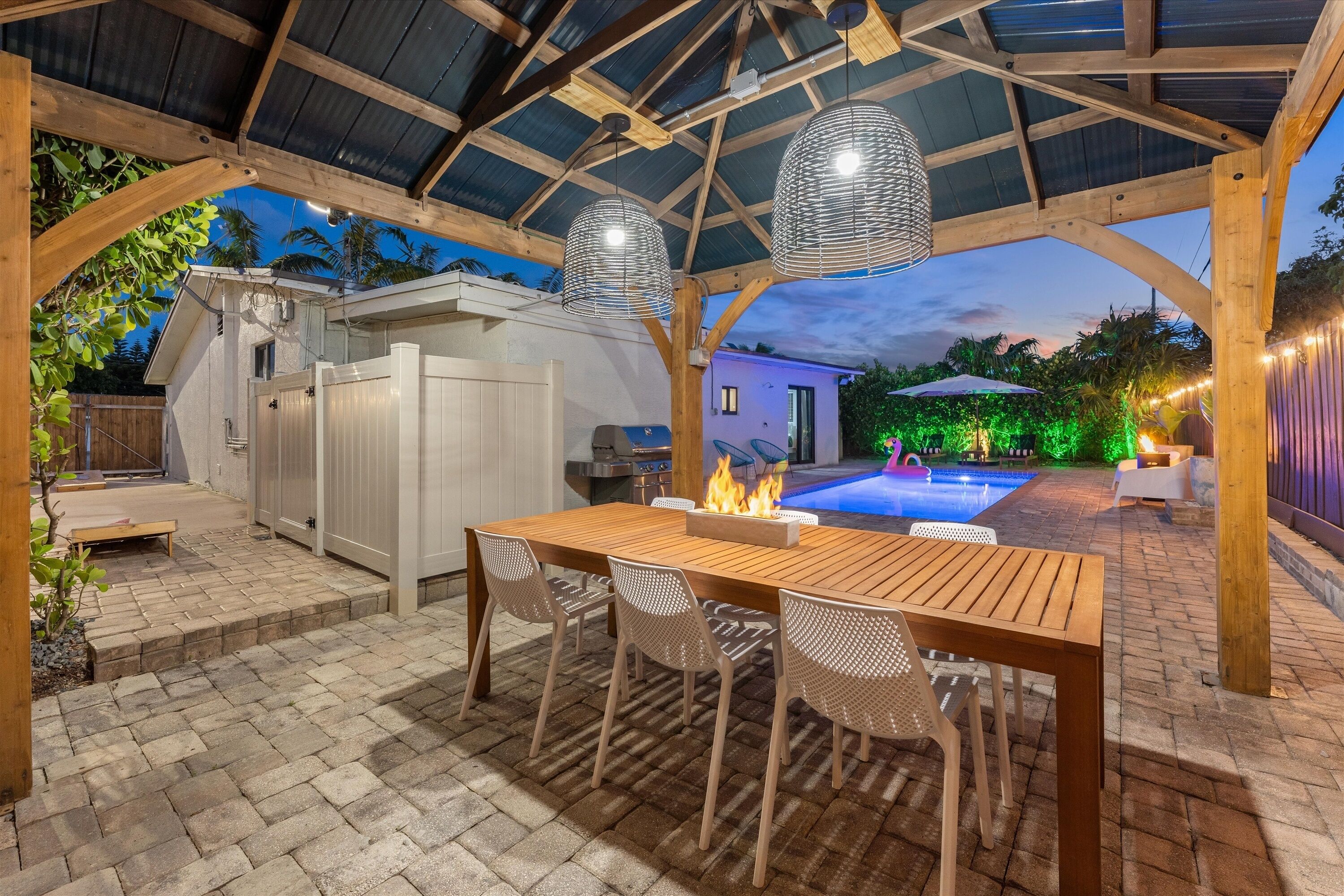 An outdoor patio with a wooden table and chairs, a barbecue grill, and a swimming pool in the background.