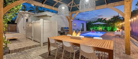 An outdoor patio with a wooden table and chairs, a barbecue grill, and a swimming pool in the background.