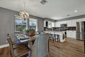 A modern kitchen with white cabinets, stainless steel appliances, and a dining area with wicker chairs and a chandelier. - Weitz Haven: Pool, Hot-tub, More! (Fredericksburg)