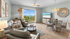 A living room with a sofa, chairs, a TV, a dining table, and a balcony with a view of the ocean. - Beach Resort 402 - Gulf View, Zero-Entry Lagoon Pool, Hot Tub, Deeded Beach! (Miramar Beach)