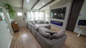 A modern living room with a long grey sectional sofa, a flat-screen TV mounted on the wall, and a wooden floor. - Family & Pet Friendly Home in Boca Raton w fenced backyard, jacuzzi & playground (Boca Raton)