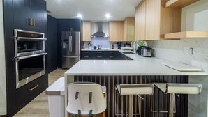 A modern kitchen with dark cabinetry, stainless steel appliances, a central island, and bar stools. - Family & Pet Friendly Home in Boca Raton w fenced backyard, jacuzzi & playground (Boca Raton)