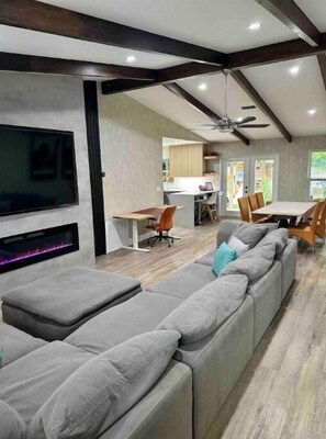 Un salon moderne avec un grand canapé d’angle gris, un téléviseur à écran plat fixé au mur et un ventilateur de plafond.