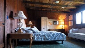 Ein gemütliches Schlafzimmer mit Holzbalkendecke, einem Bett mit gemusterten Betttextilien, einem Nachttisch mit Lampe, einer Kommode und einem Fenster mit Tageslicht.