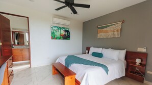 A bedroom with a bed, a bench, a ceiling fan, a wall-mounted air conditioner, a painting, and a macrame wall hanging. - King Suite in Playa Coyote, King Suite just steps from pool with swim up bar! (San Francisco de Coyote)