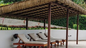 A covered outdoor seating area with lounge chairs and a thatched roof. - King Suite in Playa Coyote, King Suite just steps from pool with swim up bar! (San Francisco de Coyote)