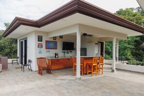A covered outdoor dining area with a kitchen in the background, wooden furniture, and a view of the surrounding greenery.