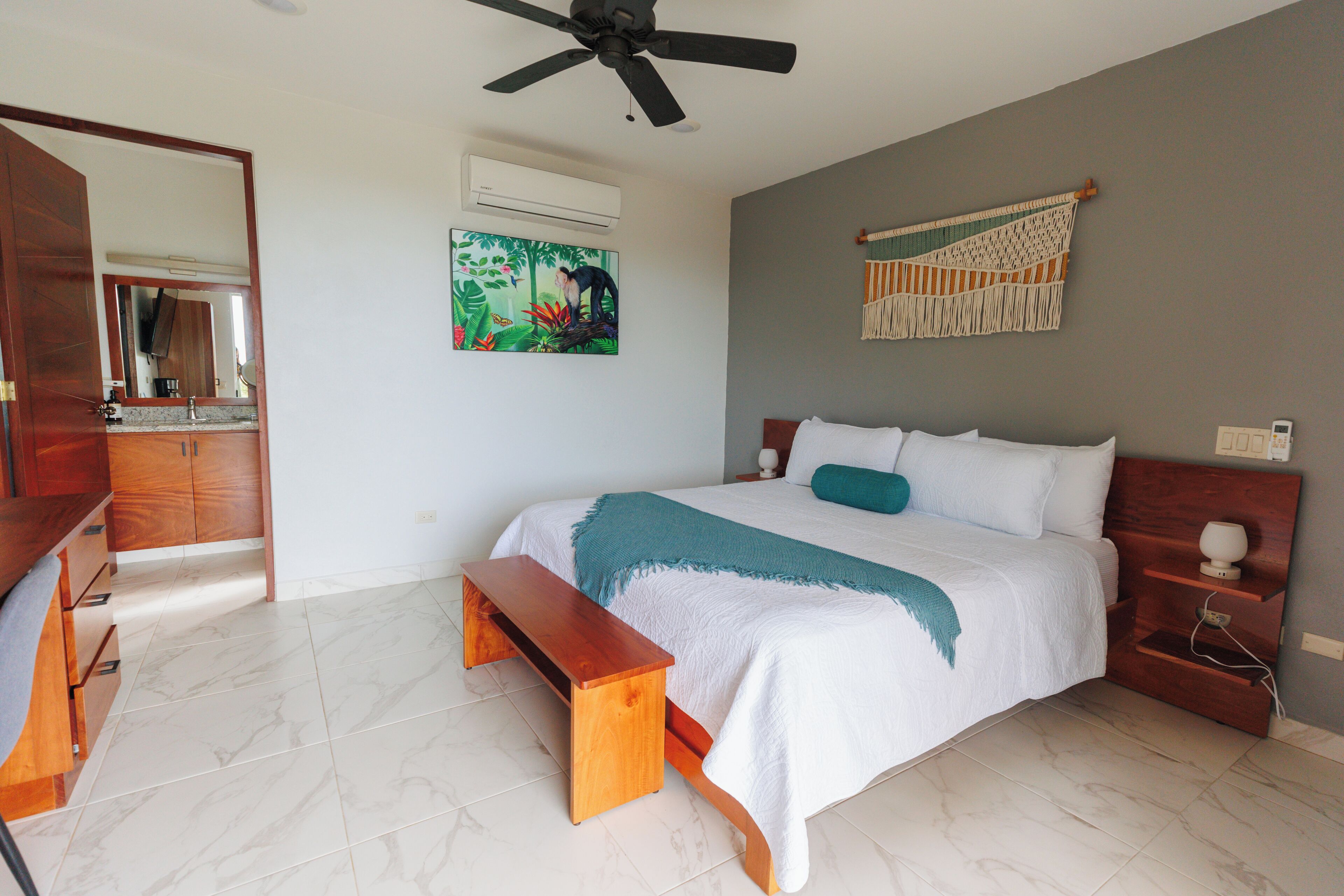 Une chambre à coucher comprenant un lit, un banc, un ventilateur de plafond, un climatiseur mural, un tableau et une décoration murale en macramé.