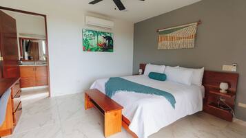 Une chambre à coucher comprenant un lit, un banc, un ventilateur de plafond, un climatiseur mural, un tableau et une décoration murale en macramé.