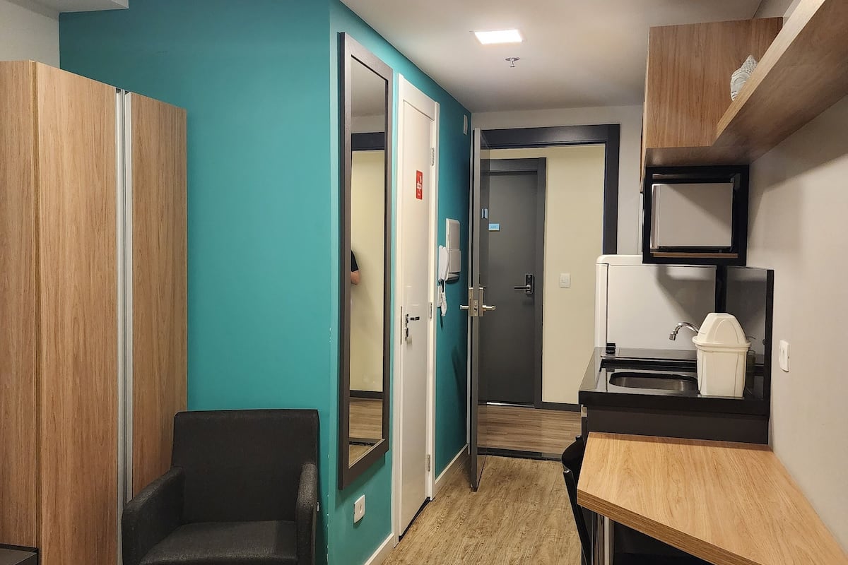 A hotel room with a bed, a desk, a chair, a wardrobe, a mirror, a door, and a kitchenette.