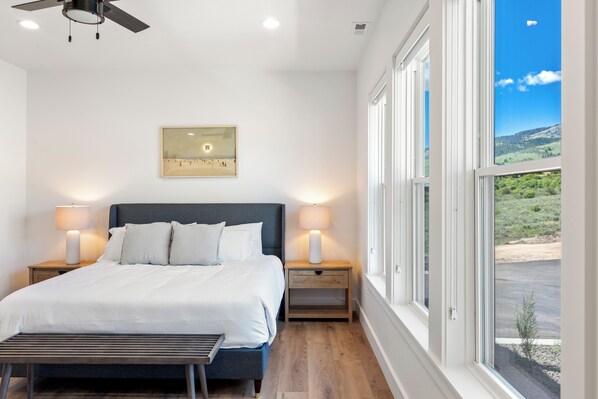 Une chambre à coucher comprenant un lit, des lampes de chevet, une table de nuit, un banc, un ventilateur de plafond et une grande fenêtre donnant sur un paysage.