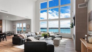 En rummelig stue med et stort vindue der giver udsigt til havet, et moderne sofasæt og et spiseområde med bord og stole.