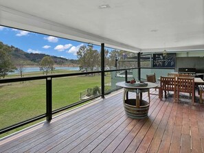 A balcony with a view of a lake and hills, a round table with a drink, and wooden chairs.