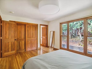 Un dormitorio con suelo de madera, una cama grande, una ventana con vistas a árboles y una puerta que da a otra habitación.