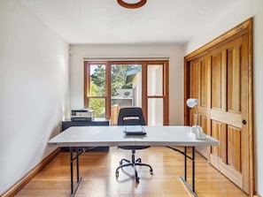 A room with a large table, a chair, a desk with a computer, and a door with a view to the outside.