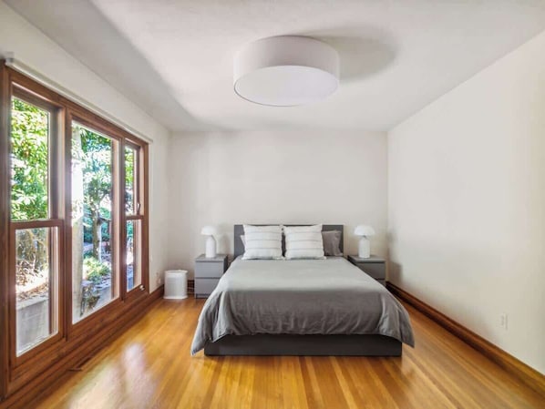 A bedroom with a large bed, two bedside tables, a ceiling light, and a view of greenery through large windows.