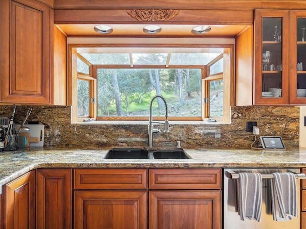 A kitchen with wooden cabinets, granite countertops, a double sink, and a window with a view of trees.