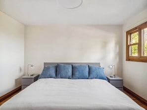 A bedroom with a large bed, two bedside tables, and a window with a view of greenery.