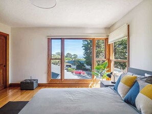 A bedroom with a large bed, a nightstand, a lamp, and a view of a street and trees through a sliding glass door.