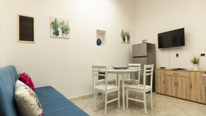 Un espacio habitable compacto con un sofá, una mesa de comedor con sillas, un refrigerador, un televisor montado en la pared y un mueble pequeño.