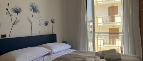 A neatly made bed with white linens and rolled towels, a blue headboard, and a wall with floral wall art.