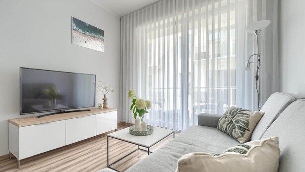 A modern living room with a flat-screen TV on a white cabinet, a grey sofa, a coffee table with decorative items, and sheer curtains allowing natural light.