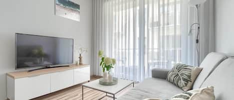 A modern living room with a flat-screen TV on a white cabinet, a grey sofa, a coffee table with decorative items, and sheer curtains allowing natural light.