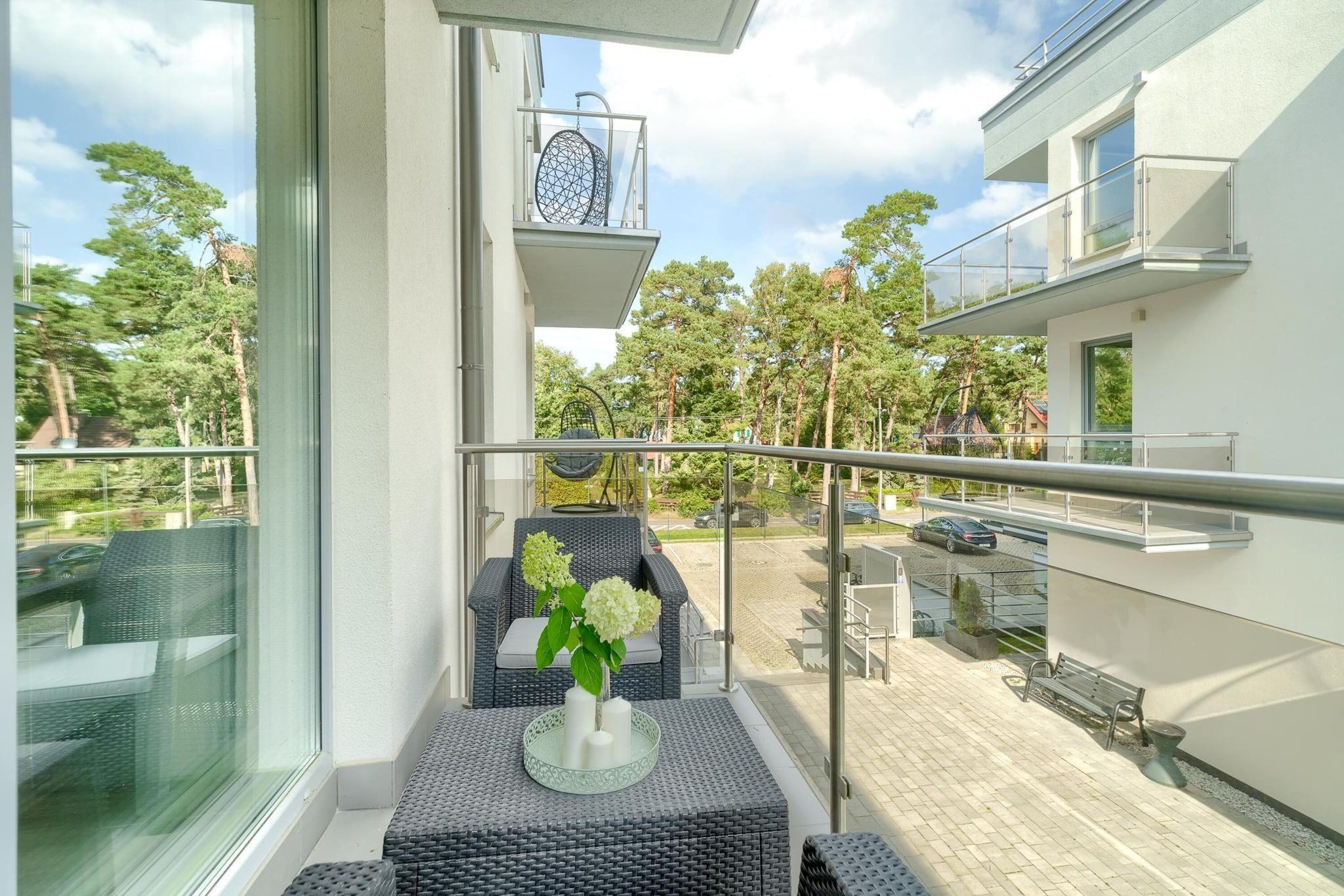 A balcony with wicker furniture, a glass table with candles, and a view of a residential area with trees and parked cars.