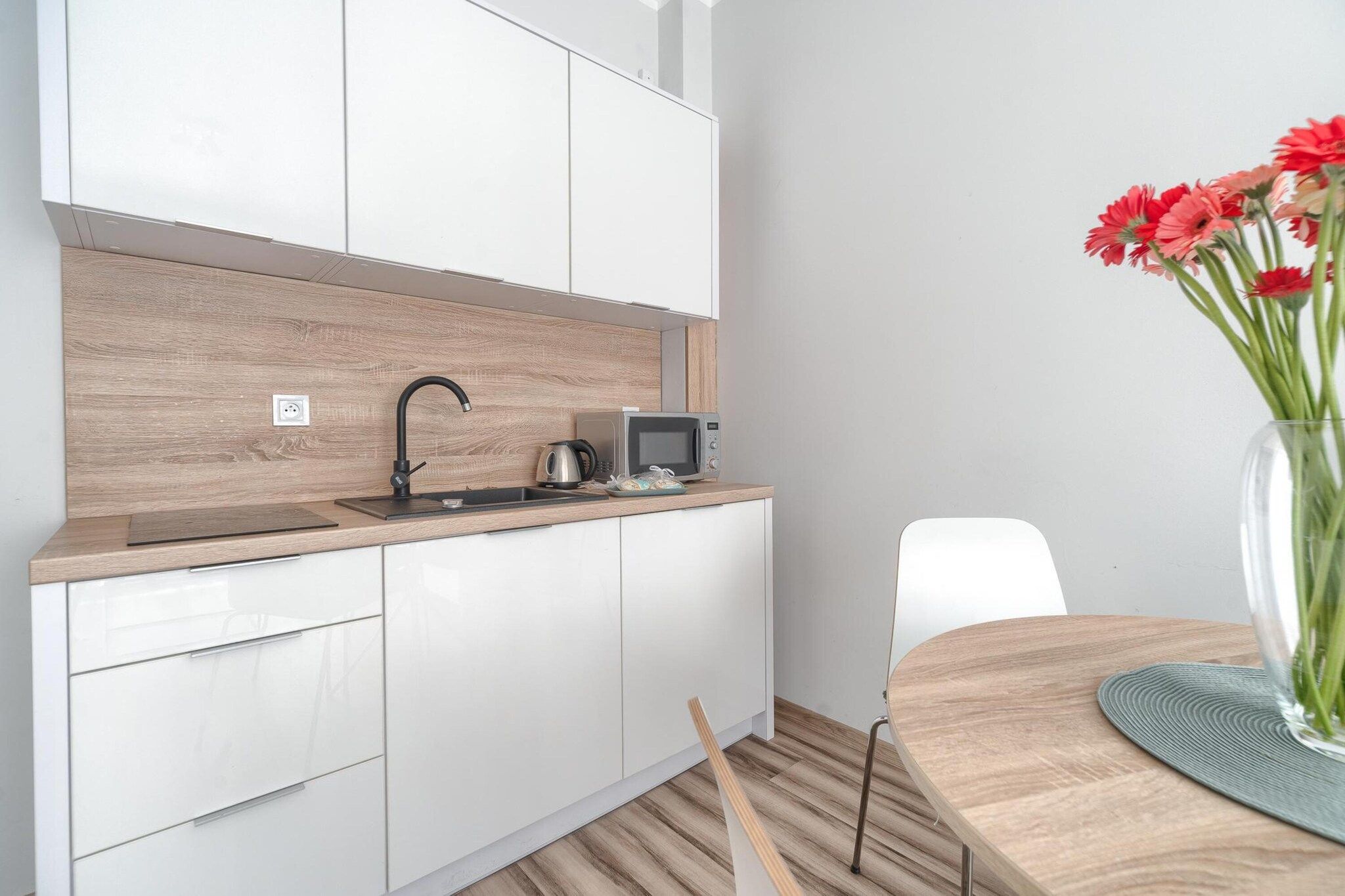 Eine moderne Küche mit weißen Schränken, einer Holzrückwand, einem schwarzen Wasserhahn, einer Mikrowelle und einem Blumenstrauß auf dem Esstisch.