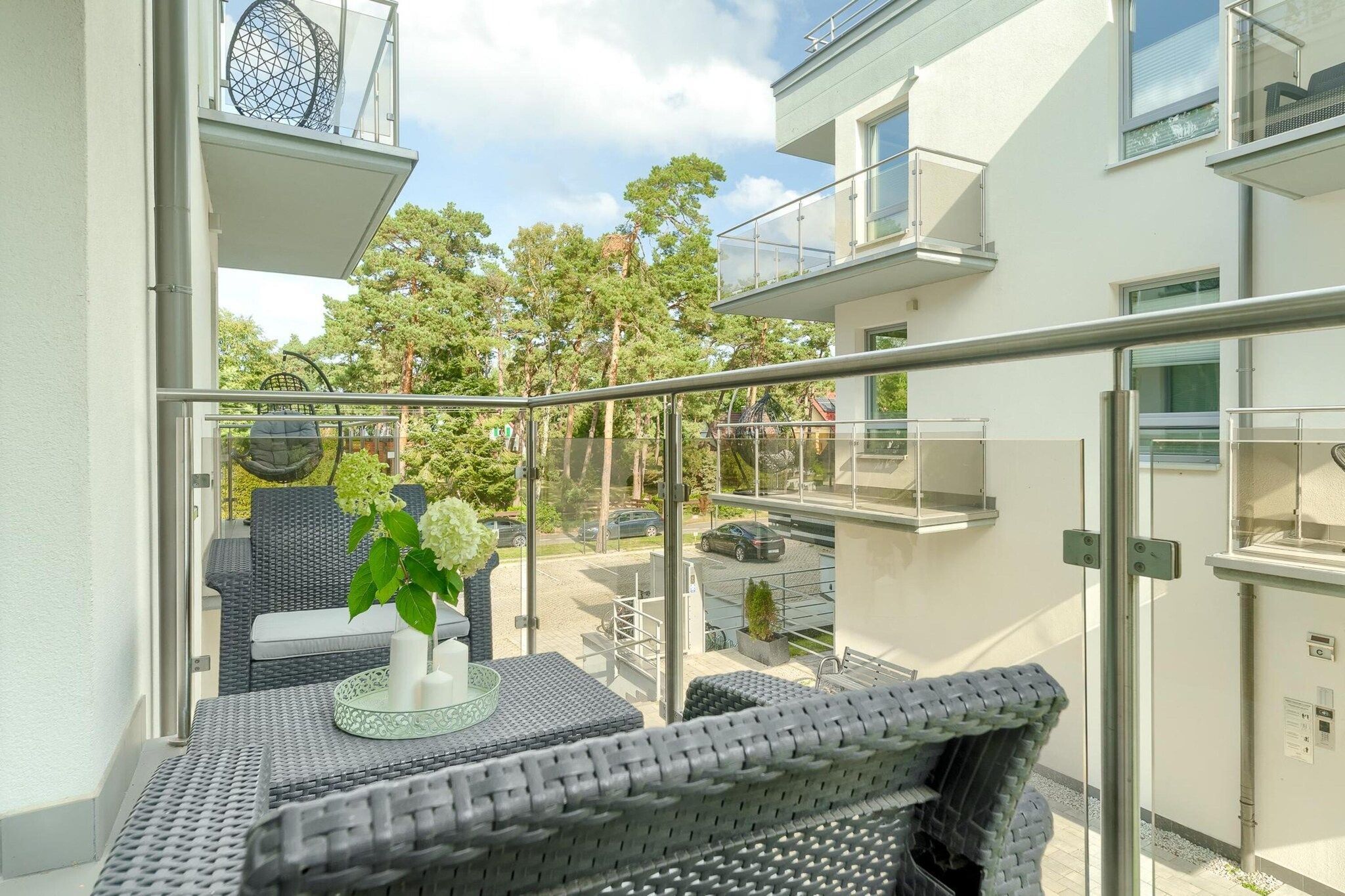 Ein Balkon mit Korbmöbeln, einer Glastrennwand und Blick auf einen Parkplatz sowie Bäume.