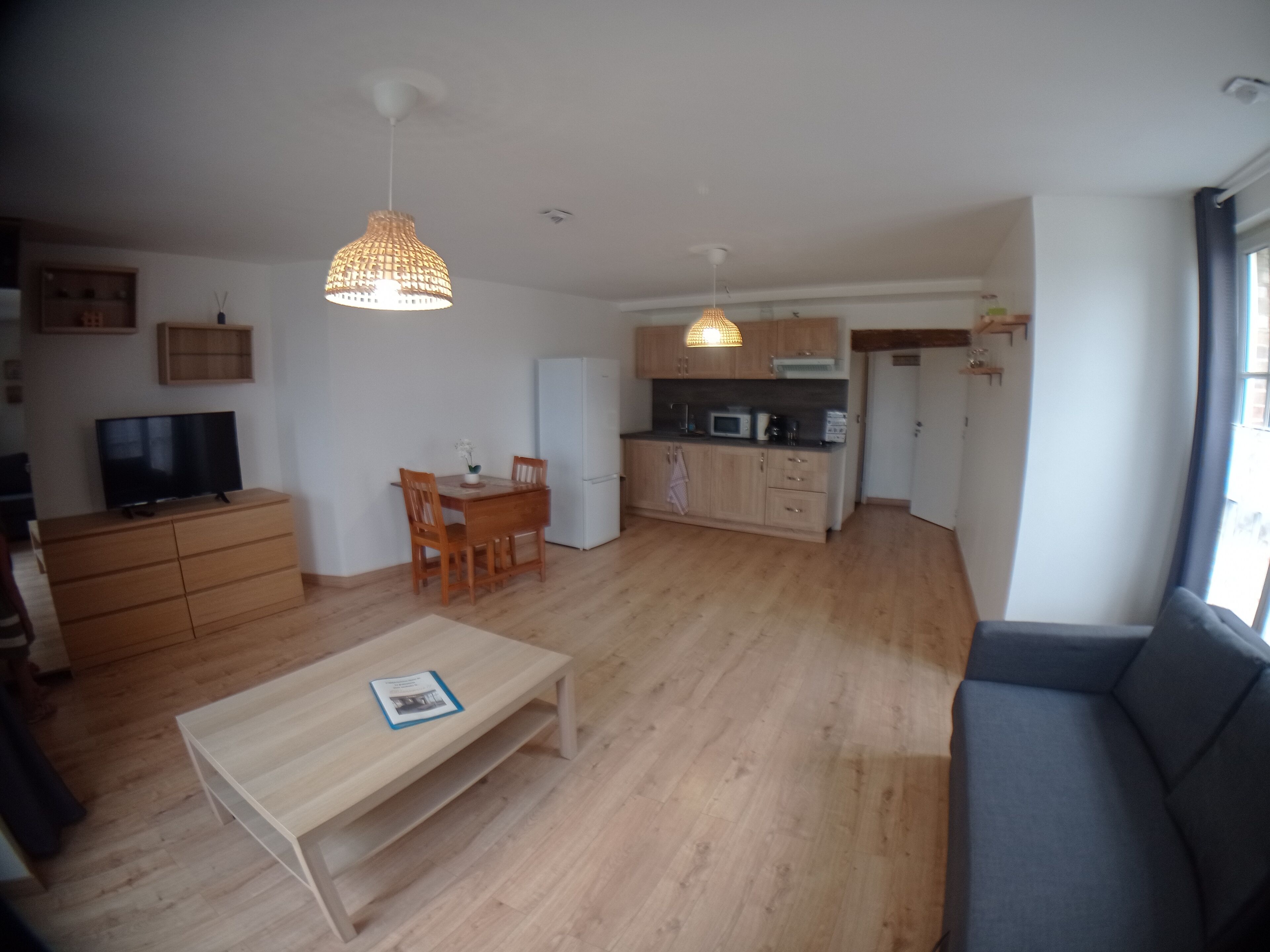 A spacious living room with a wooden floor, a dining area with a table and chairs, a kitchen with modern appliances, and a large window providing natural light.