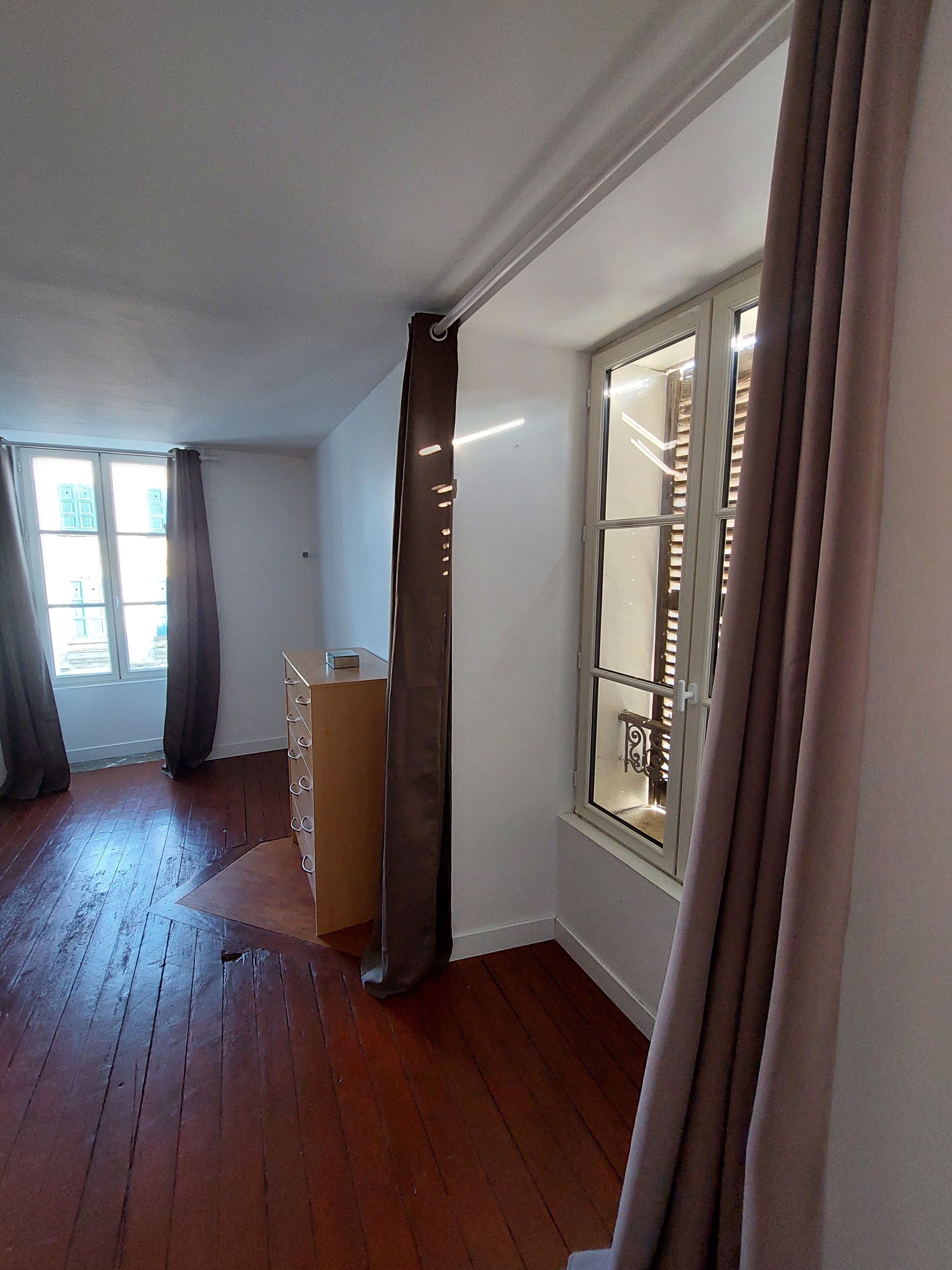 A room with wooden flooring, a radiator, a window with blinds, and a door with a curtain.