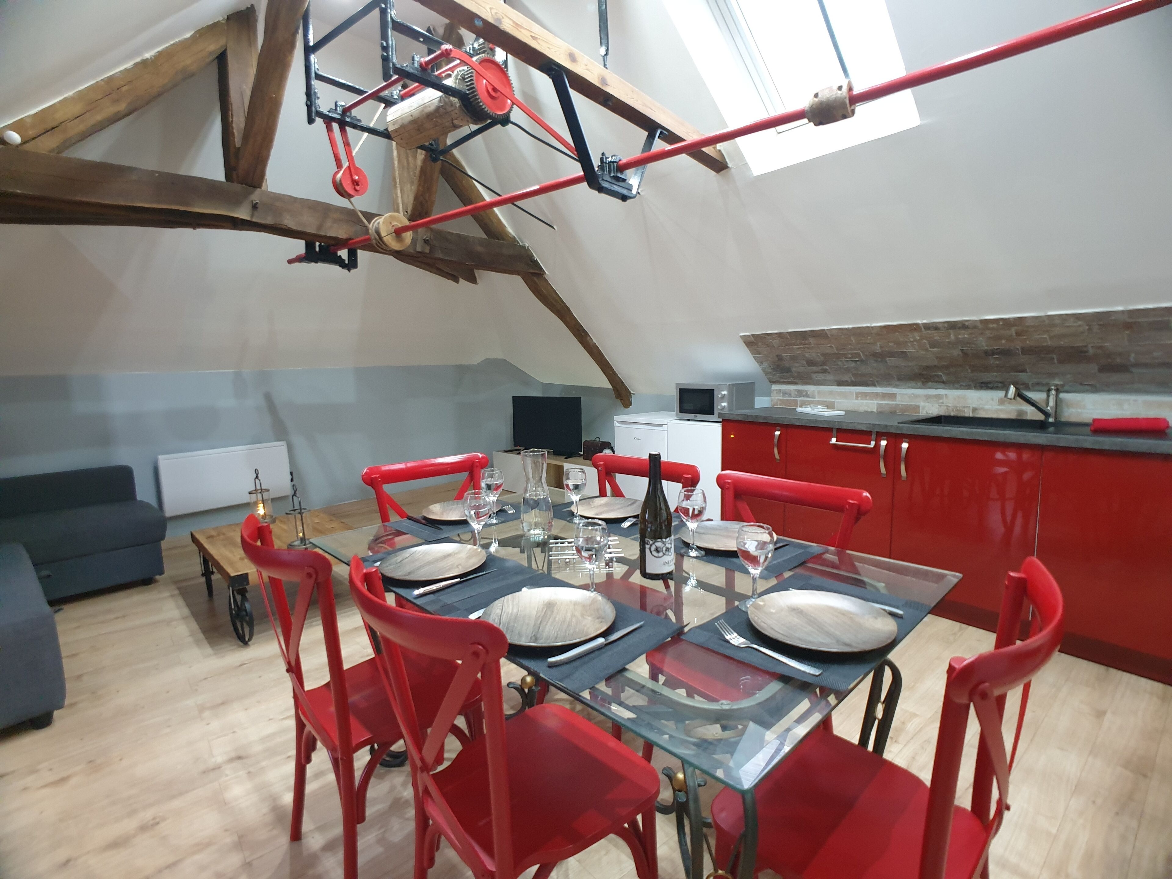 A dining area with a high ceiling, exposed wooden beams, and red chairs. A table is set with plates, glasses, and a bottle.