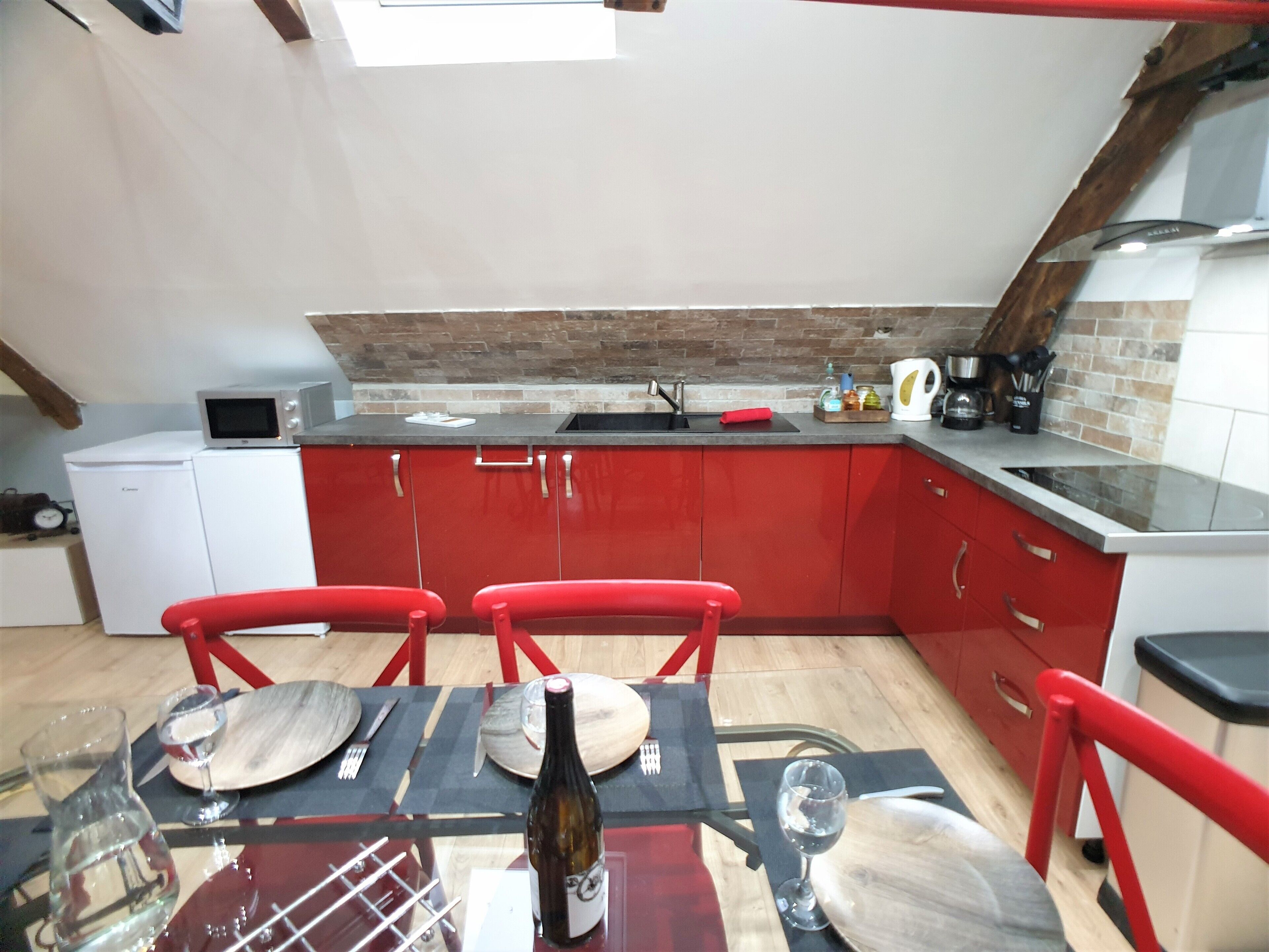 Een kleine keuken met rode kastjes, een magnetron en een spoelbak. Er staat een eettafel met stoelen en een fles wijn.