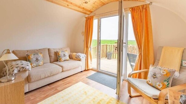 Een gezellige kamer met een houten plafond, een bank met bloemenkussens, een houten stoel en een glazen schuifdeur die leidt naar een buitenruimte.