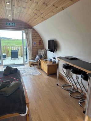 A room with a bed, a wooden desk, a television, a chair, and a view of a green landscape.