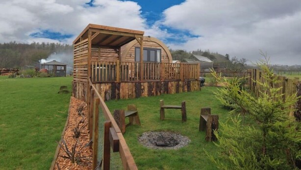Une cabane en bois dotée d’une terrasse, entourée d’une cour clôturée avec un foyer et des bancs.
