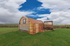 A small, single-story wooden structure with a sloped roof, a window, and a chimney, situated in a grassy field with a clear sky.