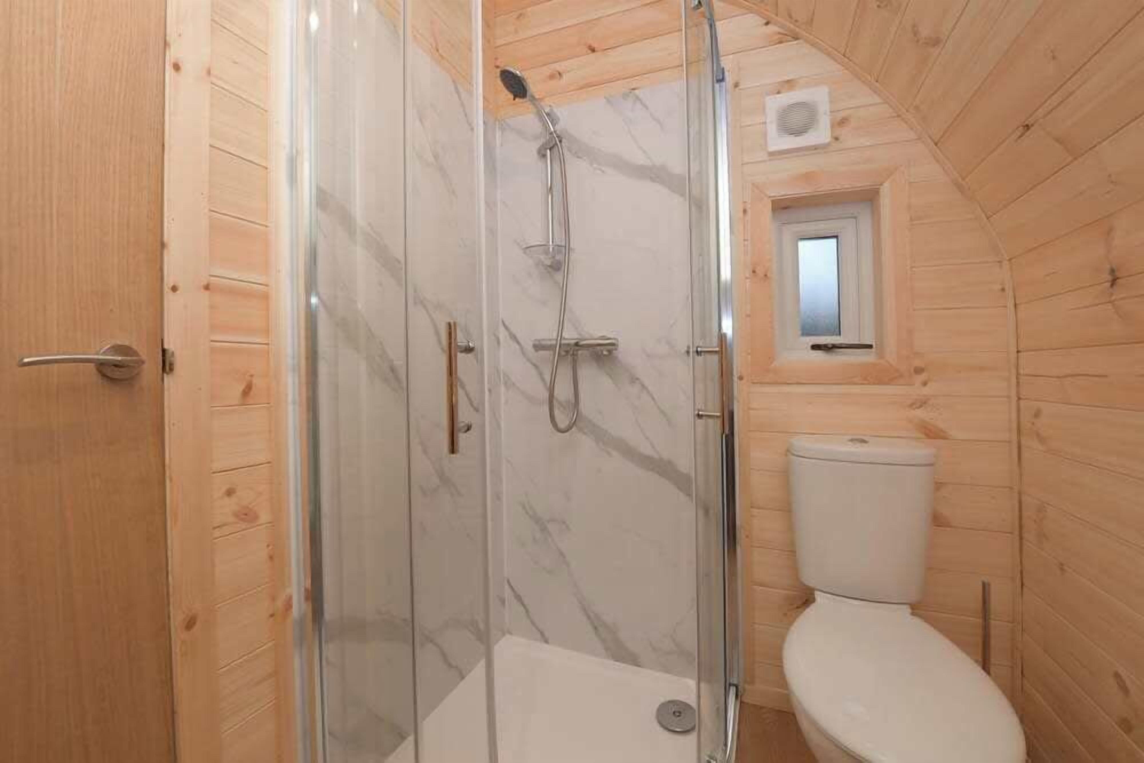 A wooden cabin bathroom with a glass-enclosed shower, a toilet, and a small window.