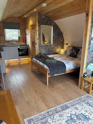 A cozy bedroom with a bed, a bedside table, a mirror, and a wooden ceiling.