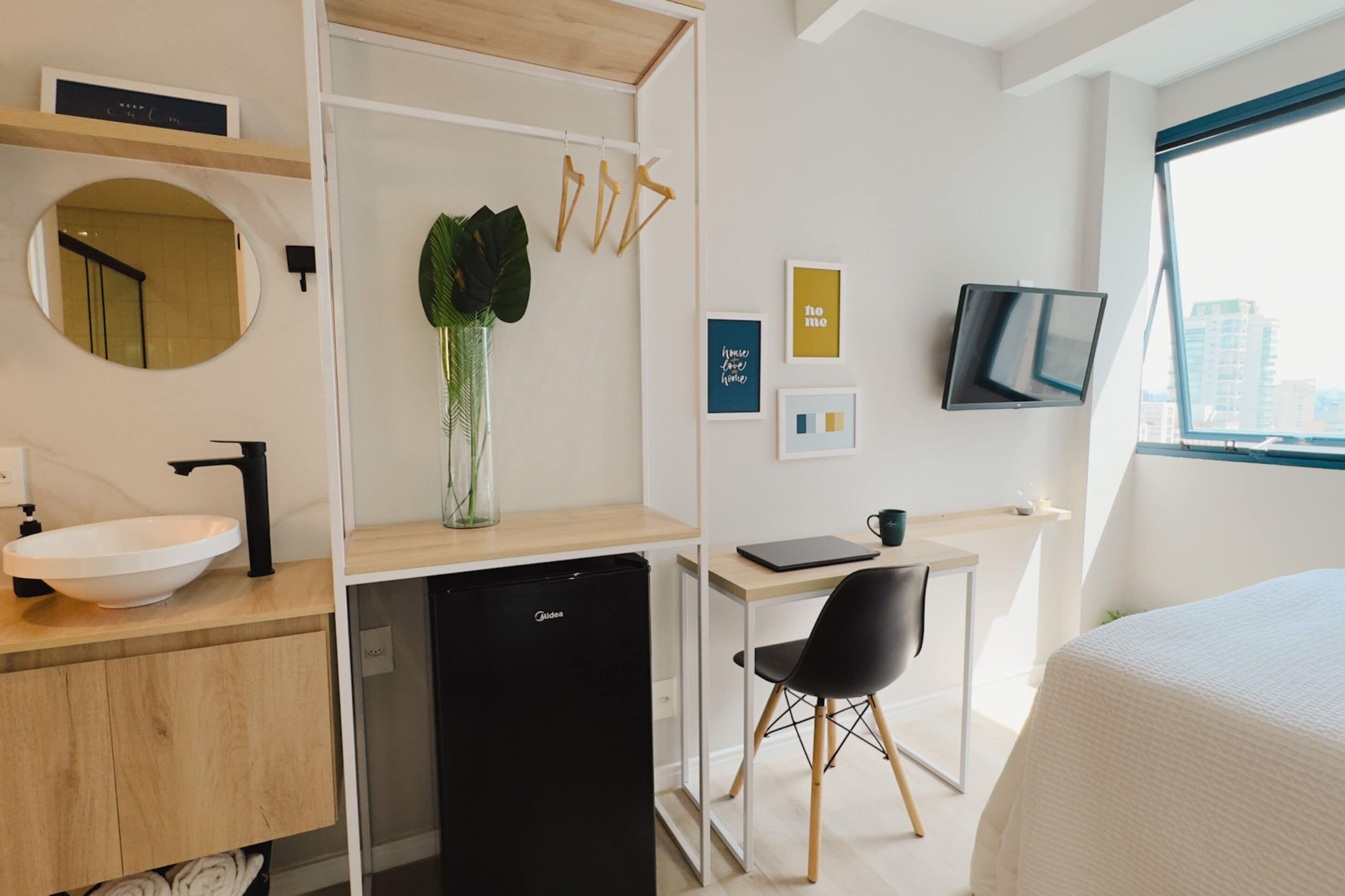 A modern hotel room with a wooden desk, a black chair, a bed, a round mirror, a black refrigerator, and a large window with a city view.