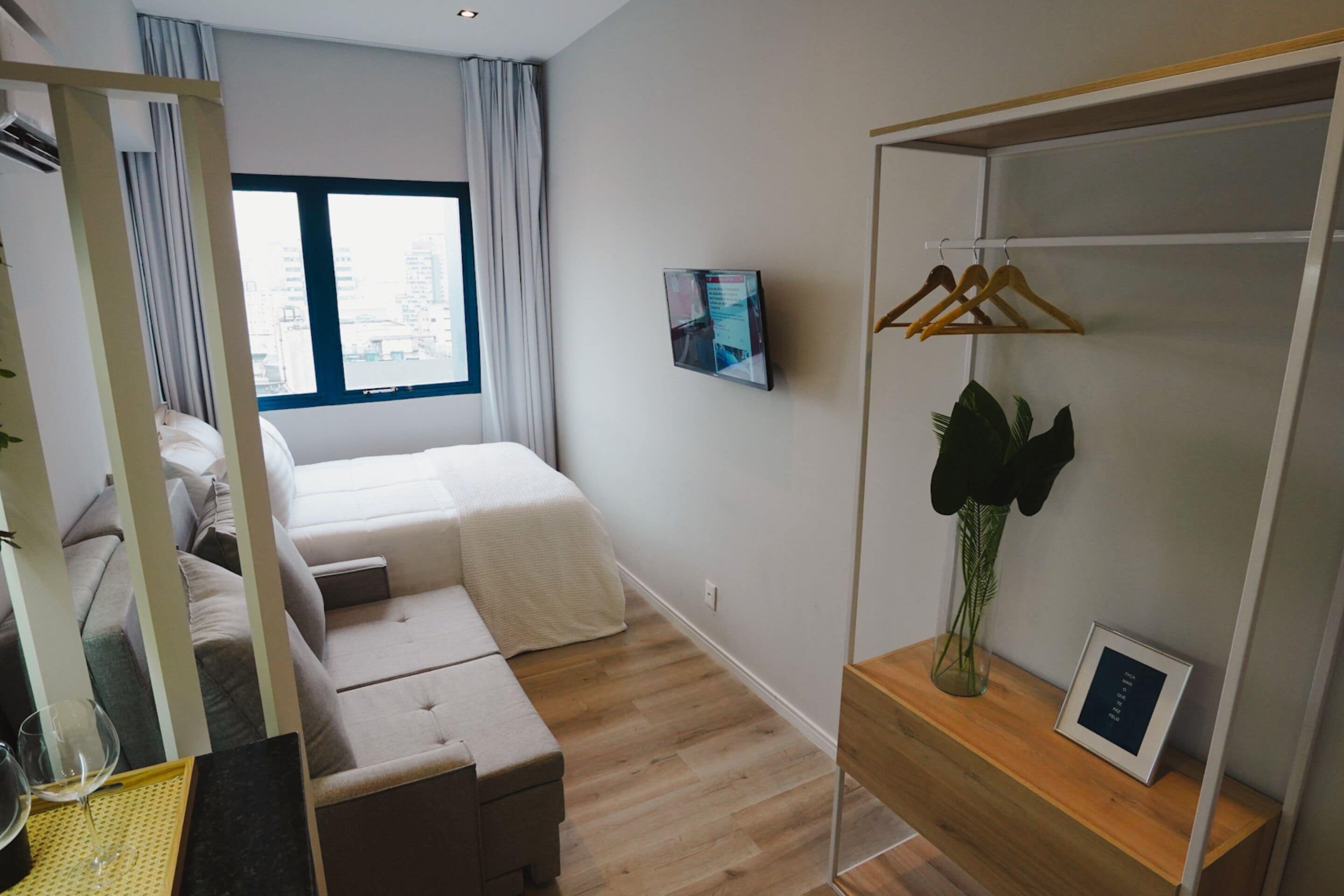 A modern hotel room with a bed, a desk, a mirror, a TV, a wardrobe, and a window with a city view.