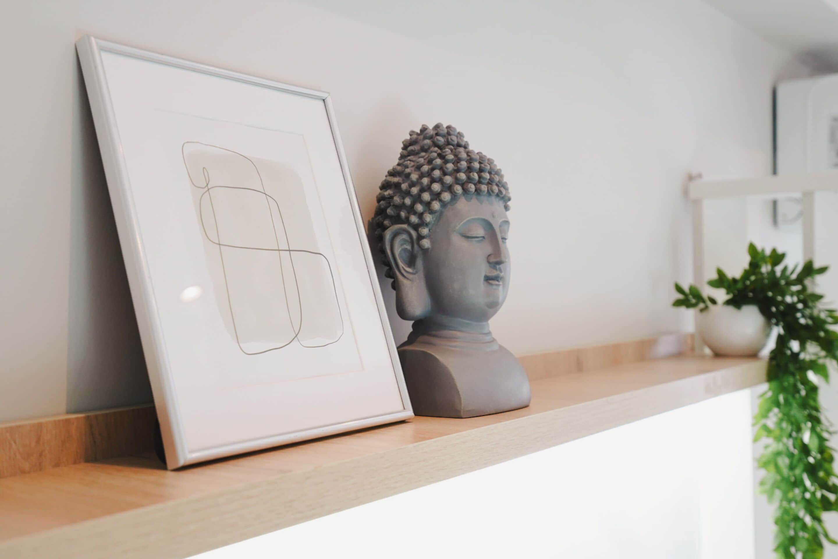 Un tableau encadré appuyé contre un mur, une statue de Bouddha et une plante en pot sur une étagère.