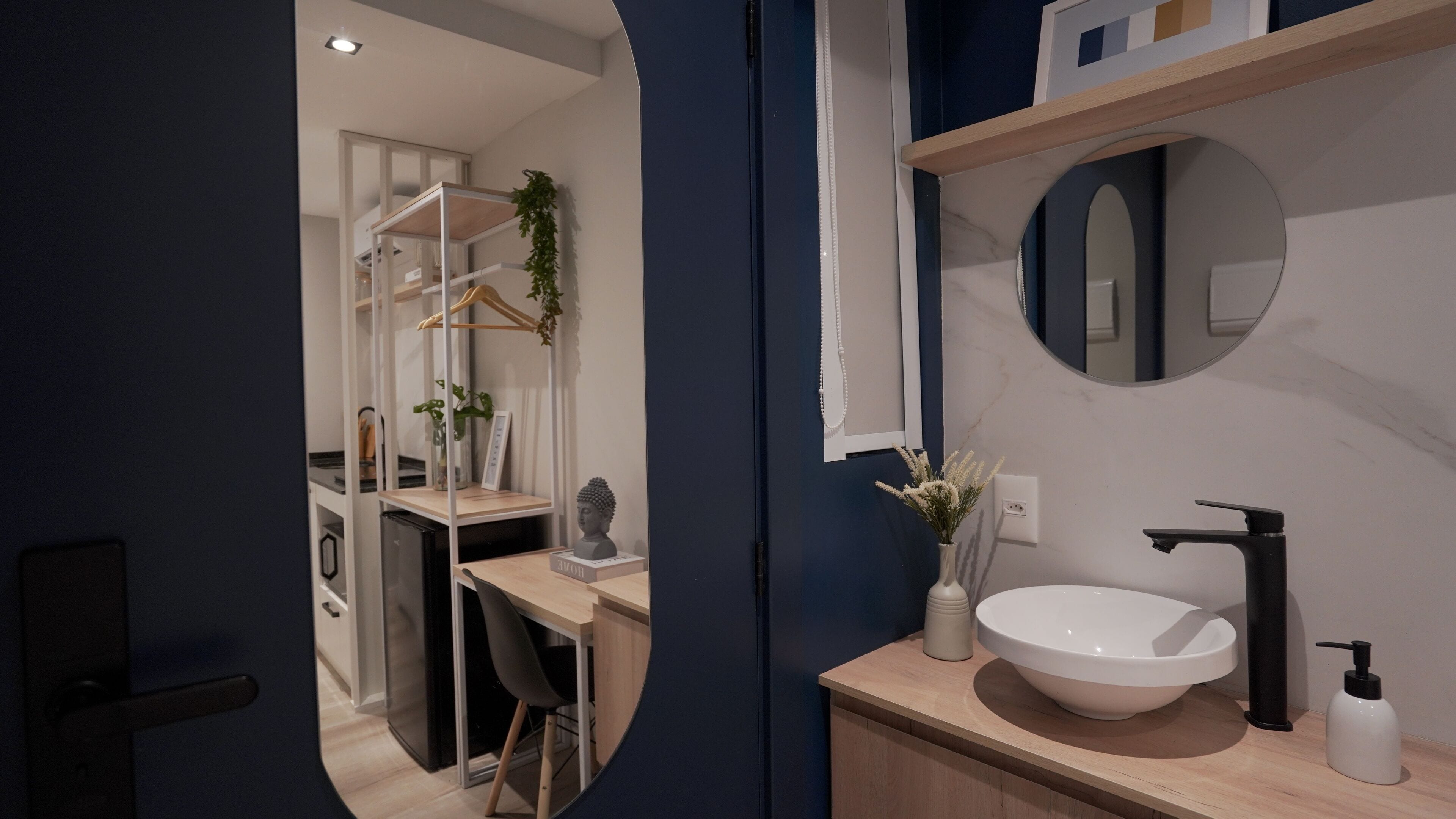 A modern bathroom with a large mirror, a white sink, and a black faucet. There is a wooden desk with a chair and a plant.