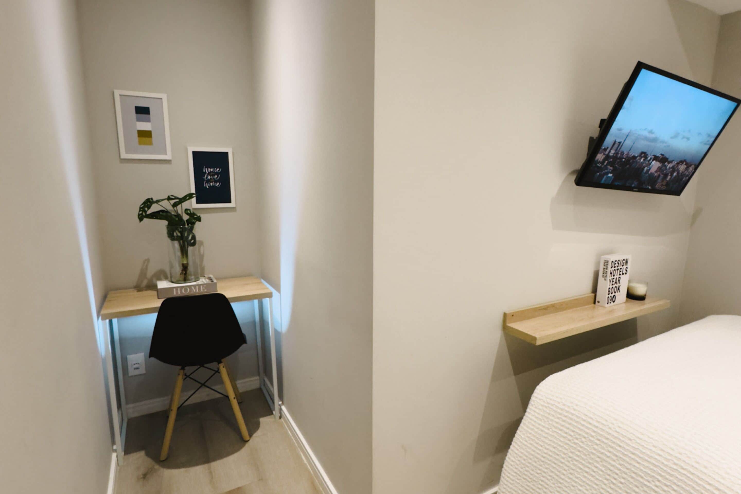 A bedroom with a bed, a desk with a chair, a TV mounted on the wall, and a shelf with a book.