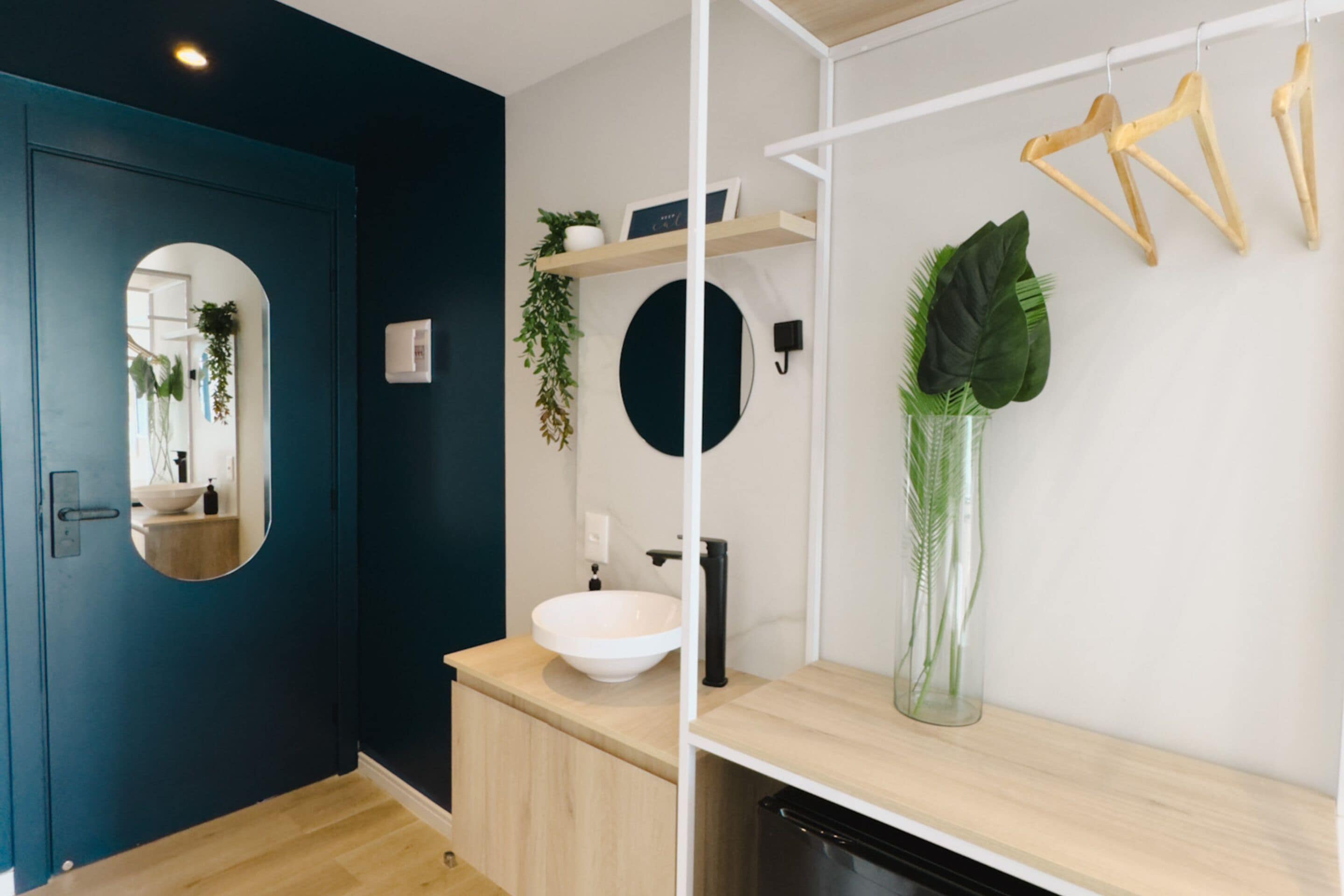 Un baño moderno con una puerta azul oscuro, un lavabo blanco, estantes de madera y un espejo.