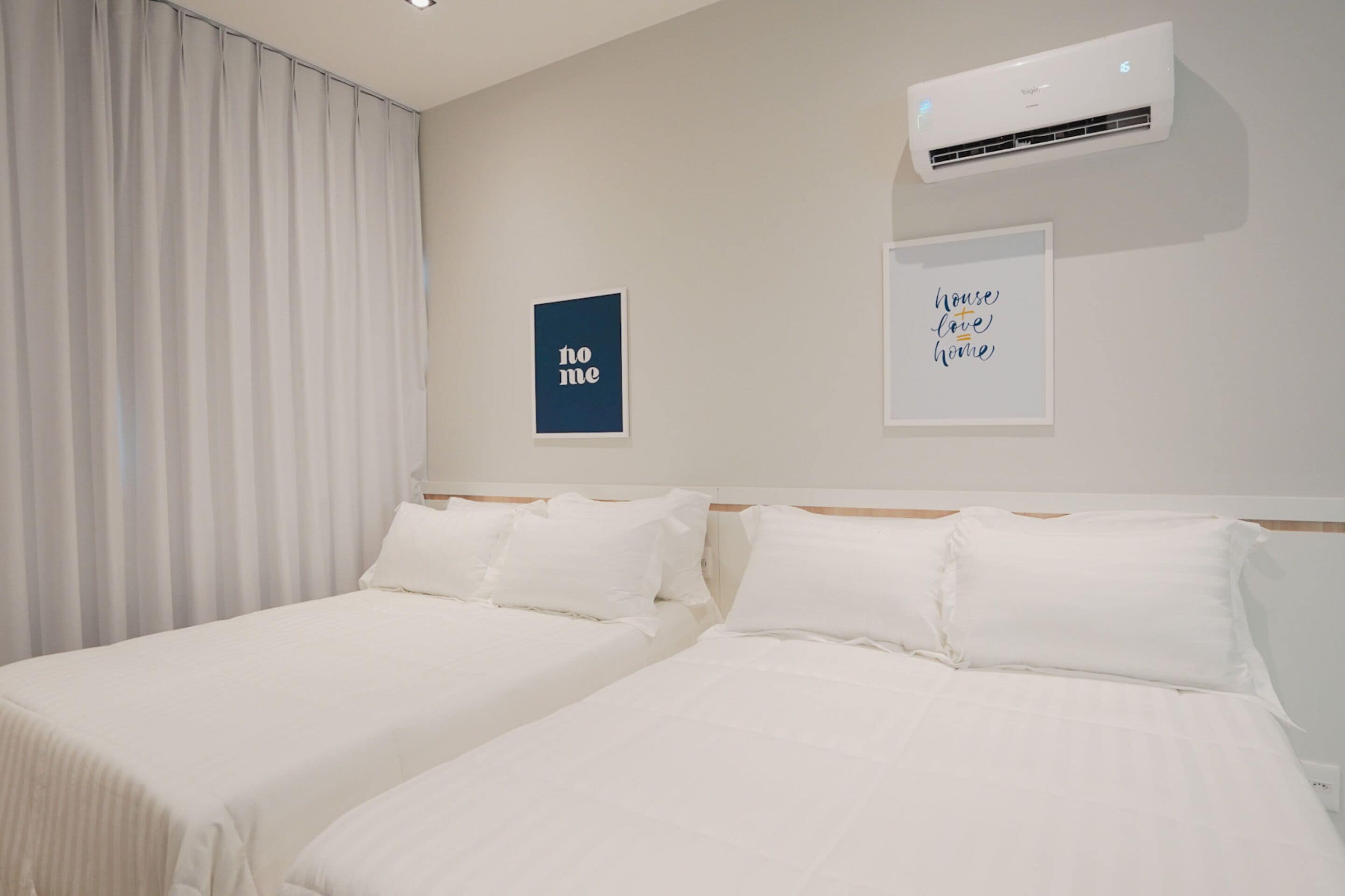 Habitación de hotel con dos camas, un ventanal con cortinas, un equipo de aire acondicionado y dos cuadros en la pared.